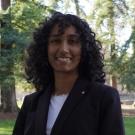 Smiling young woman with curly hair in blazer and white shirt outdoors among tall trees