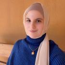 Young woman in beige hijab and blue knit sweater, soft smile, warm orange background